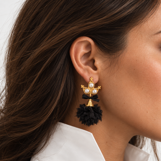A close-up side profile of a woman wearing a Tassel statement earring from Bote A Mano . The earring features a gold-toned cross-like top with white pearl-like accents and a large black tassel below, set against a clean white background.