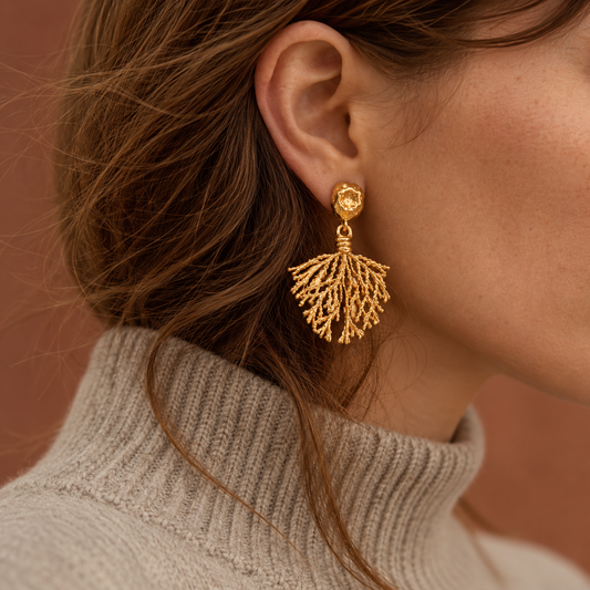 A close-up of a woman's ear wearing a gold drop earring of BOTE A MANO with a textured, fan-like coral or leafy design, paired with a beige ribbed turtleneck sweater against a warm brown background.
