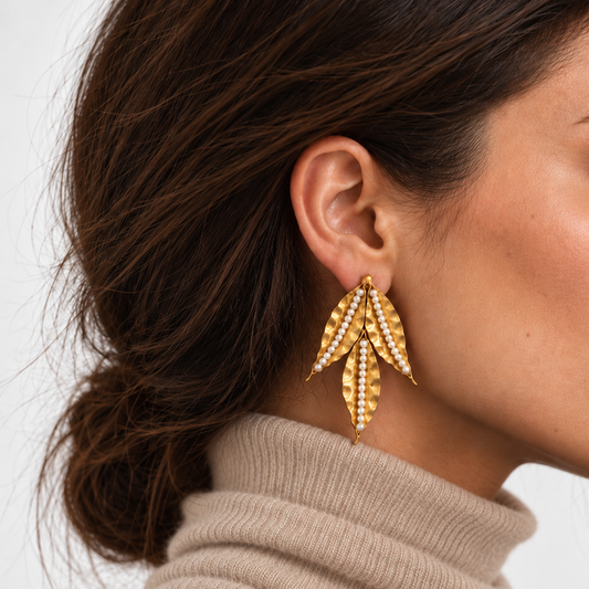 A close-up side profile of a woman wearing a large gold leaf-shaped drop earring from Bote A Mano adorned with small clear stones, paired with a beige turtleneck sweater against a light gray background.
