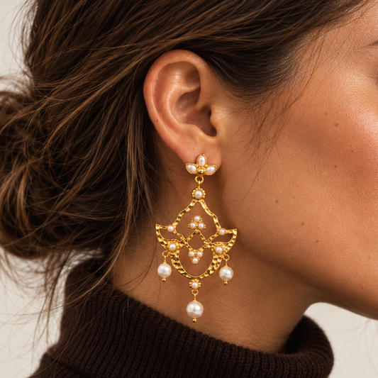 A close-up side profile of a woman wearing a large, ornate gold dangle earring adorned with small pearls and bead details from Bote A Mano, paired with a dark brown turtleneck sweater.