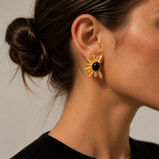 A close-up of a woman's ear wearing a gold sunburst stud Bote A Mano earring with a round black center, set against a neutral light gray background.