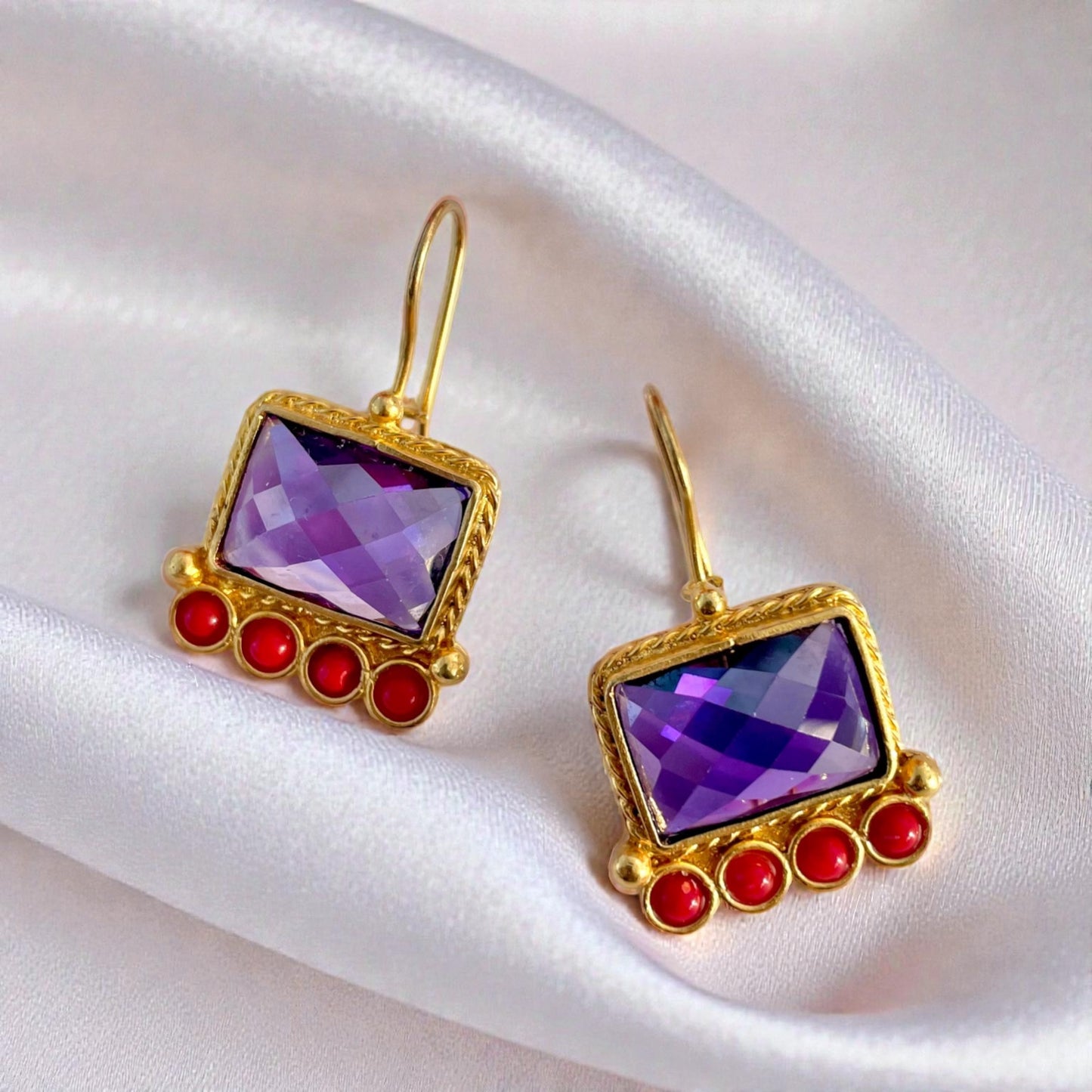 Gold earrings with purple amethyst gemstones and red accents on a white satin background