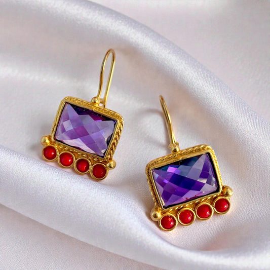 Gold earrings with purple amethyst gemstones and red accents on a white satin background