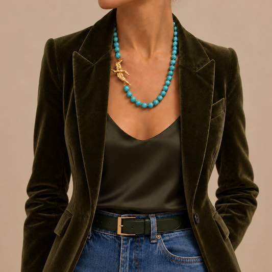 A close-up of a model wearing a dark olive green velvet blazer, a black satin camisole, blue jeans, and a chunky turquoise beaded necklace with a gold figure charm from BOTE A MANO, set against a solid beige background.