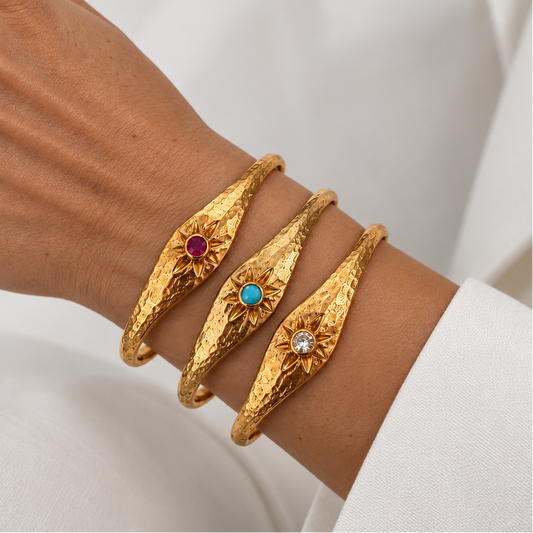 Three textured gold bangle bracelets with sunburst designs and small colored gemstone centers from Bote A Mano, worn on a person's wrist against a white fabric background.