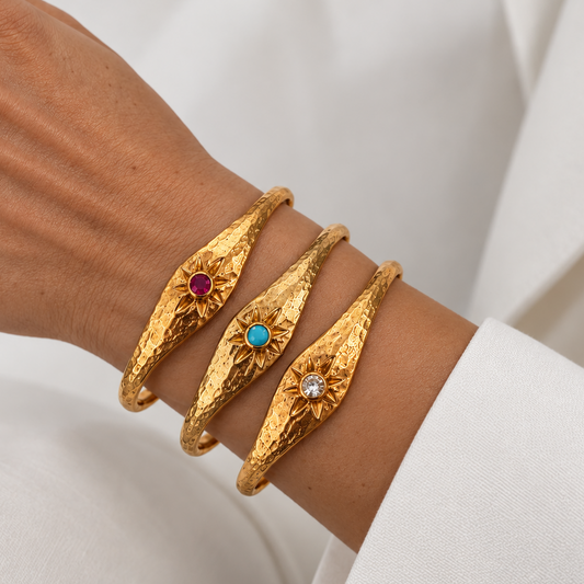 Three textured gold bangle bracelets with sunburst designs and small colored gemstone centers rom Bote A Mano, shown worn on a person's wrist against a white fabric background.