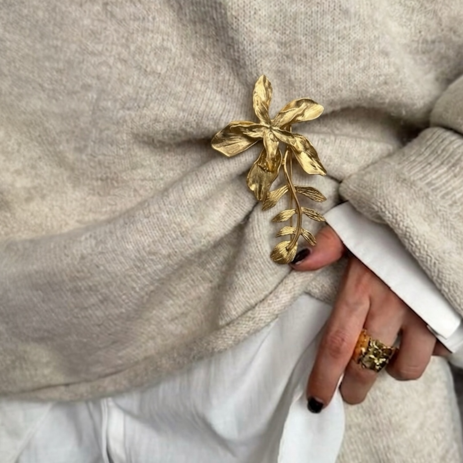 Gold leaf brooch on a beige sweater with a hand holding a white object.